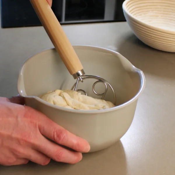 En hand håller i en ljusgrå skål med deg och använder en dansk degvisp i trä. I bakgrunden står en jäskorg i rotting på en grå köksbänk och en liten del av en svart induktionshäll.